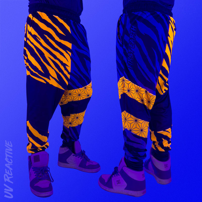 First Earth Sacred Stripes velvet harem joggers under UV blacklight on model showing glowing orange tiger stripes and geometric panels