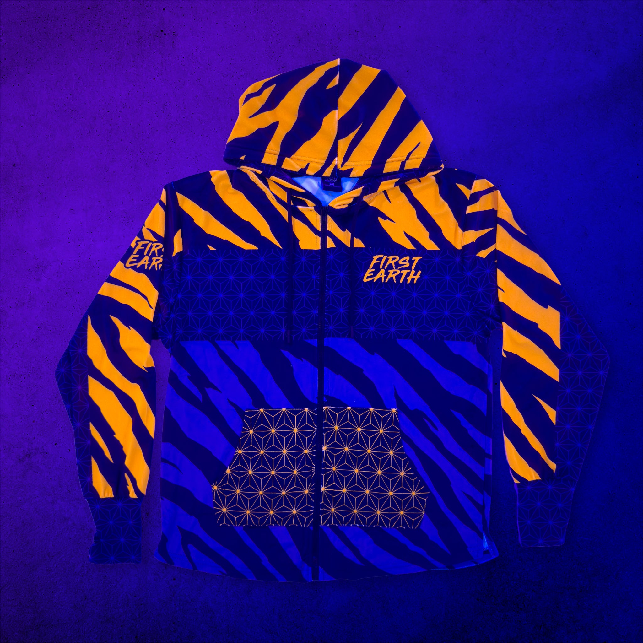 First Earth Sacred Stripes velvet hoodie glowing under UV black light with bright orange tiger stripes, geometric details, zipper, hood, and front pocket